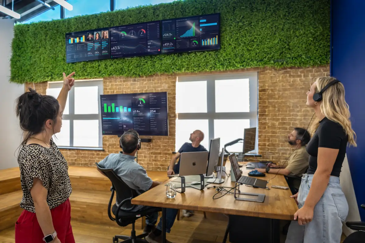 A sales team gathered around multiple TV dashboard screens showing live sales performance data