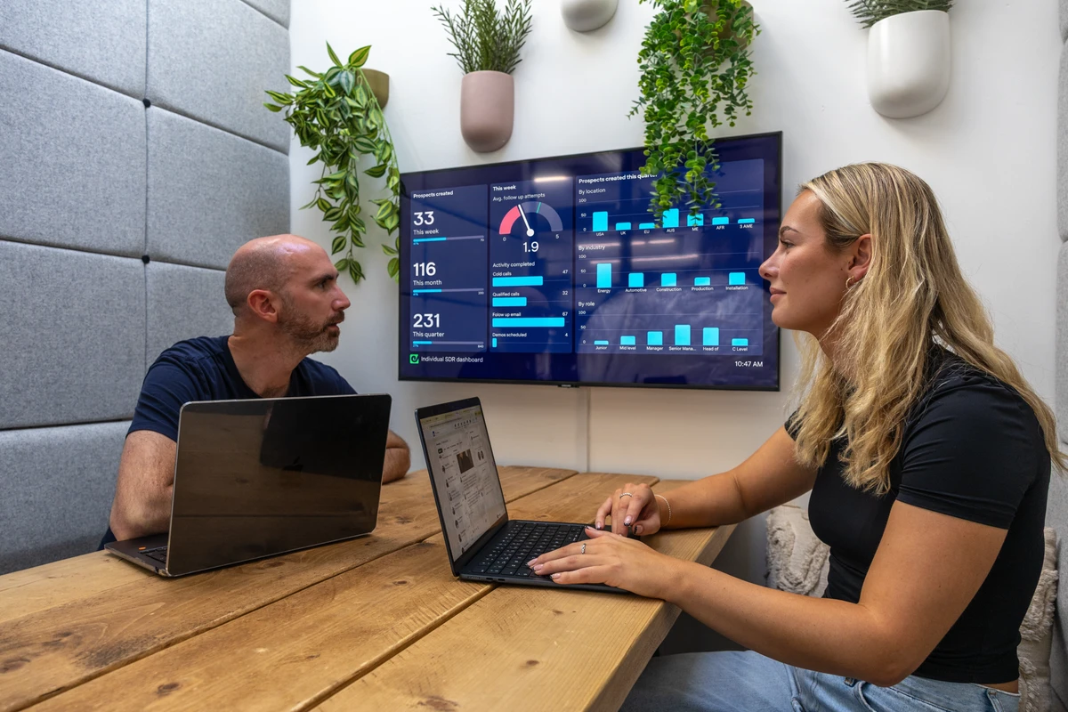 Two people in an office booth viewing a live SDR dashboard on a TV screen